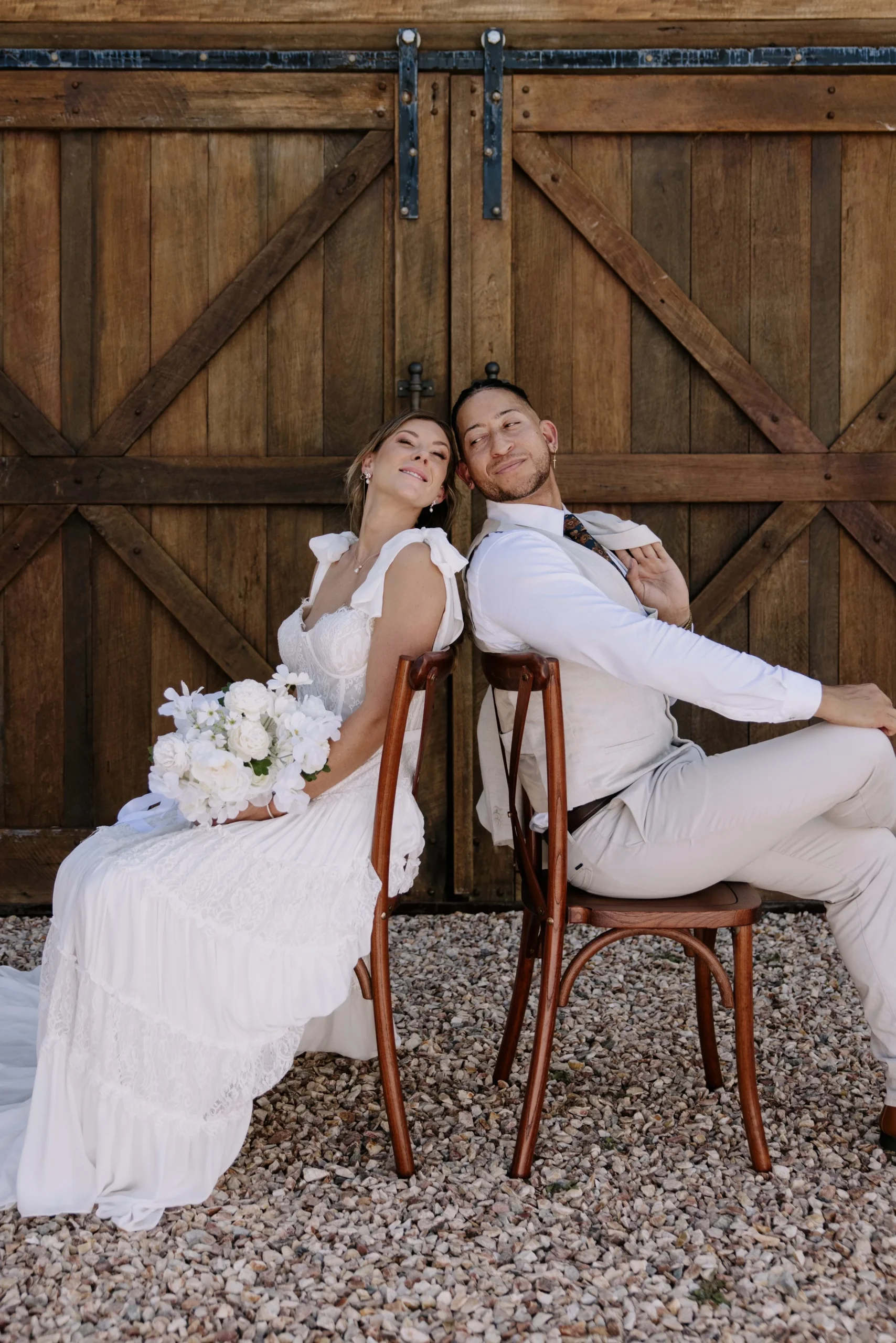Bride and groom sitting back-to-back on wooden chairs, smiling and leaning her head on his shoulder, in front of a barn door.