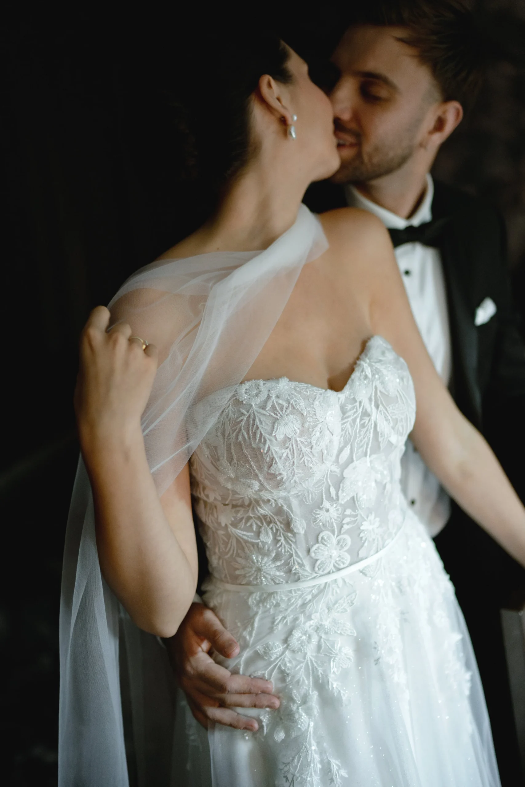 A bride and groom kissing on their wedding day, with the groom wearing a black tuxedo and the bride in a white lace wedding dress.